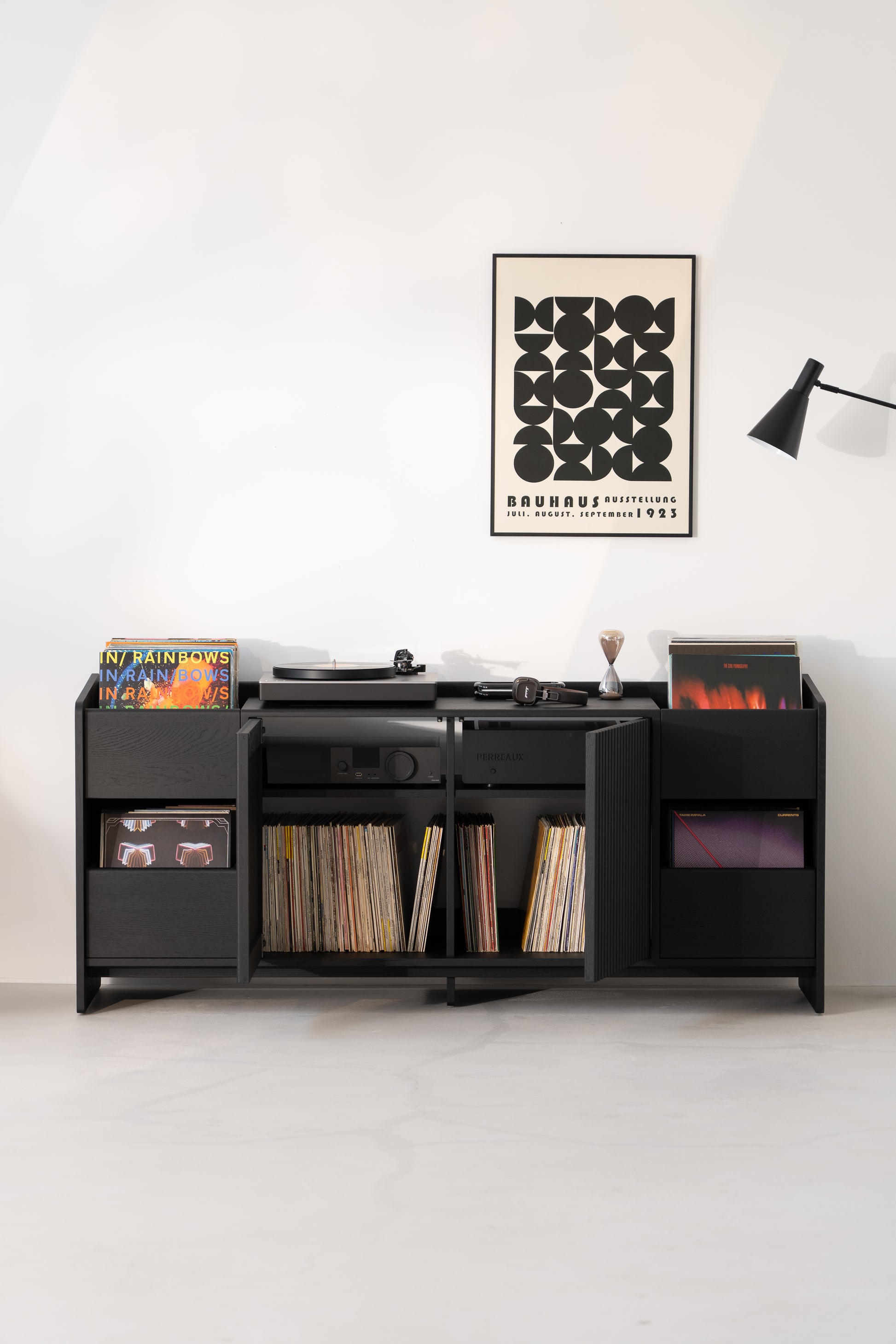 Black cabinet with records, turntable, and headphones. Bauhaus poster and lamp on white wall above.