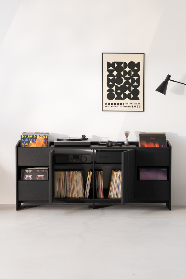 Black cabinet with records, turntable, and headphones. Bauhaus poster and lamp on white wall above.