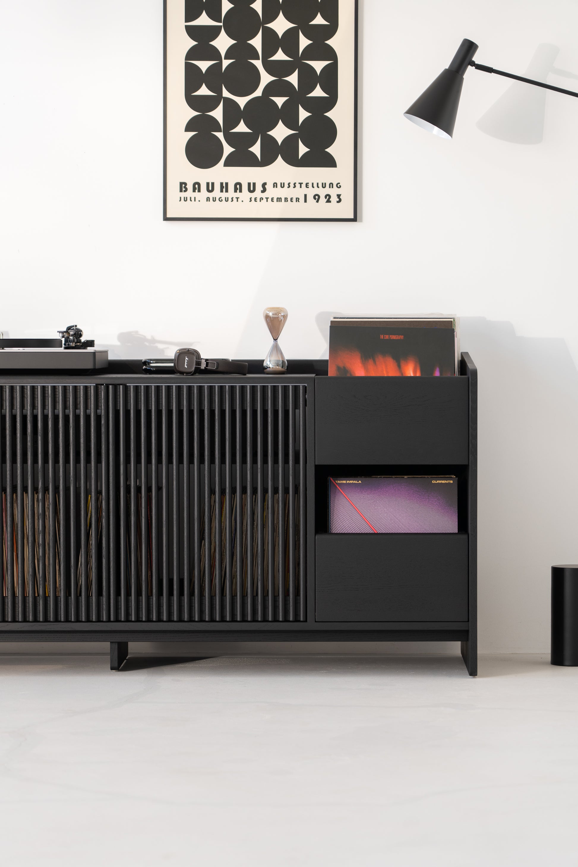 Modern black cabinet with records, books, headphones, and a Bauhaus poster above; black lamp on the side.
