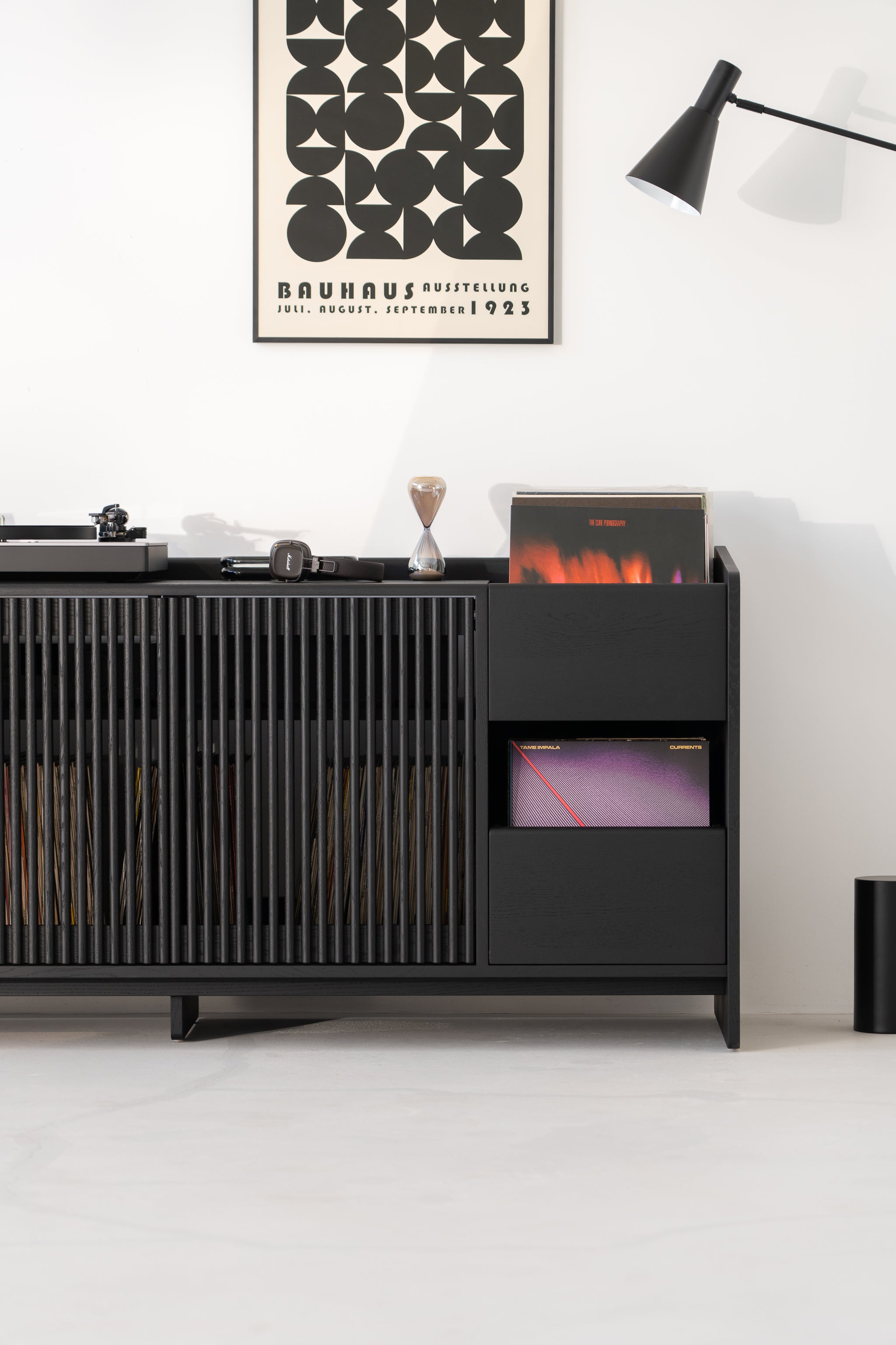 Modern black cabinet with records, books, headphones, and a Bauhaus poster above; black lamp on the side.
