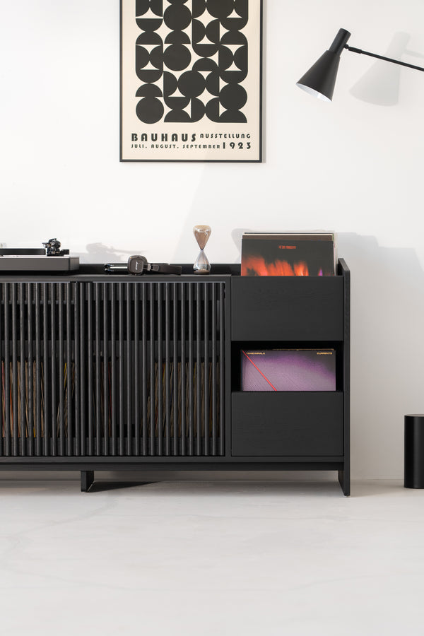 Modern black cabinet with records, books, headphones, and a Bauhaus poster above; black lamp on the side.