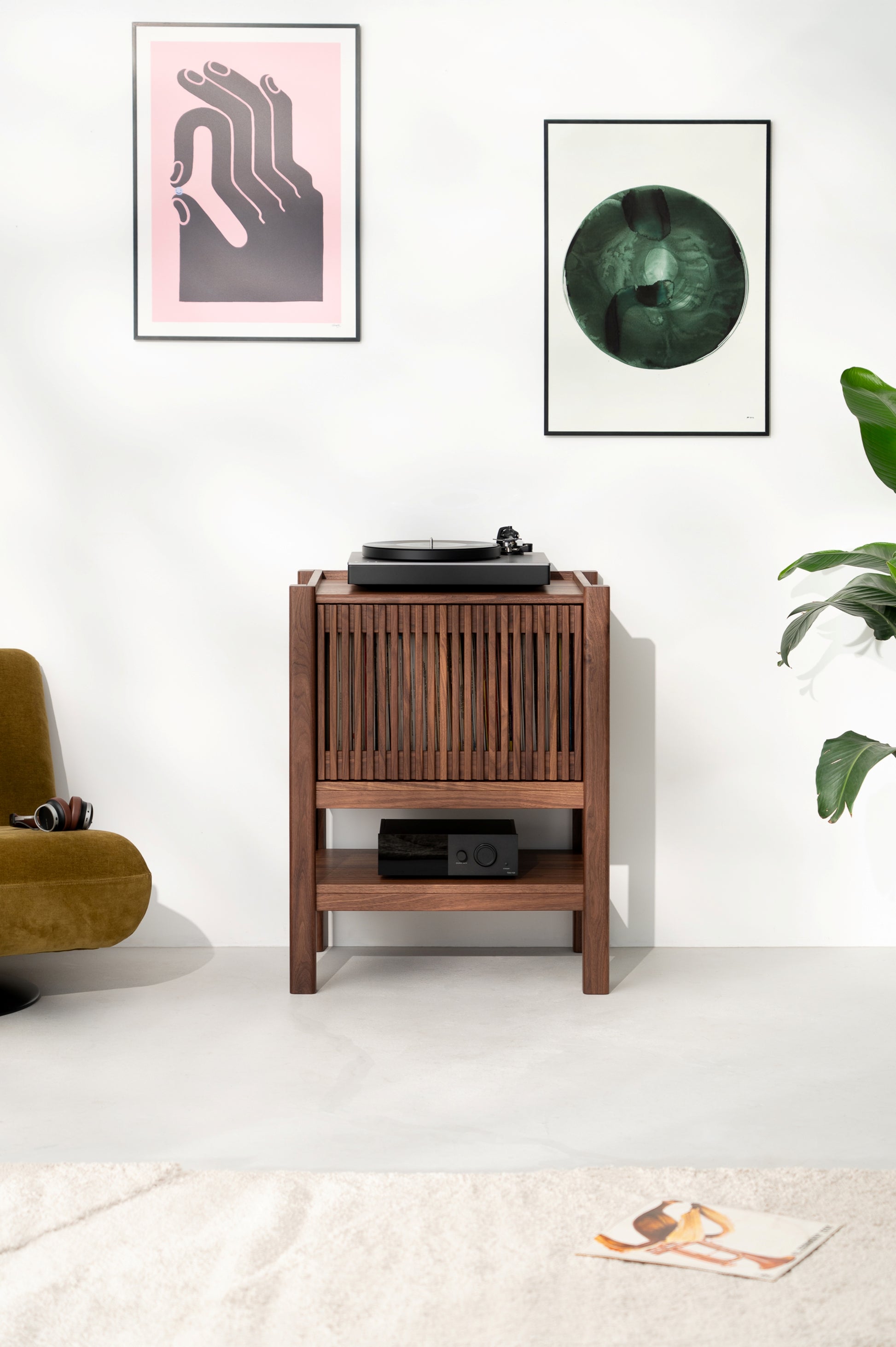 Wooden record player stand with vinyl, flanked by two framed abstract artworks, plant, and a chair with headphones.