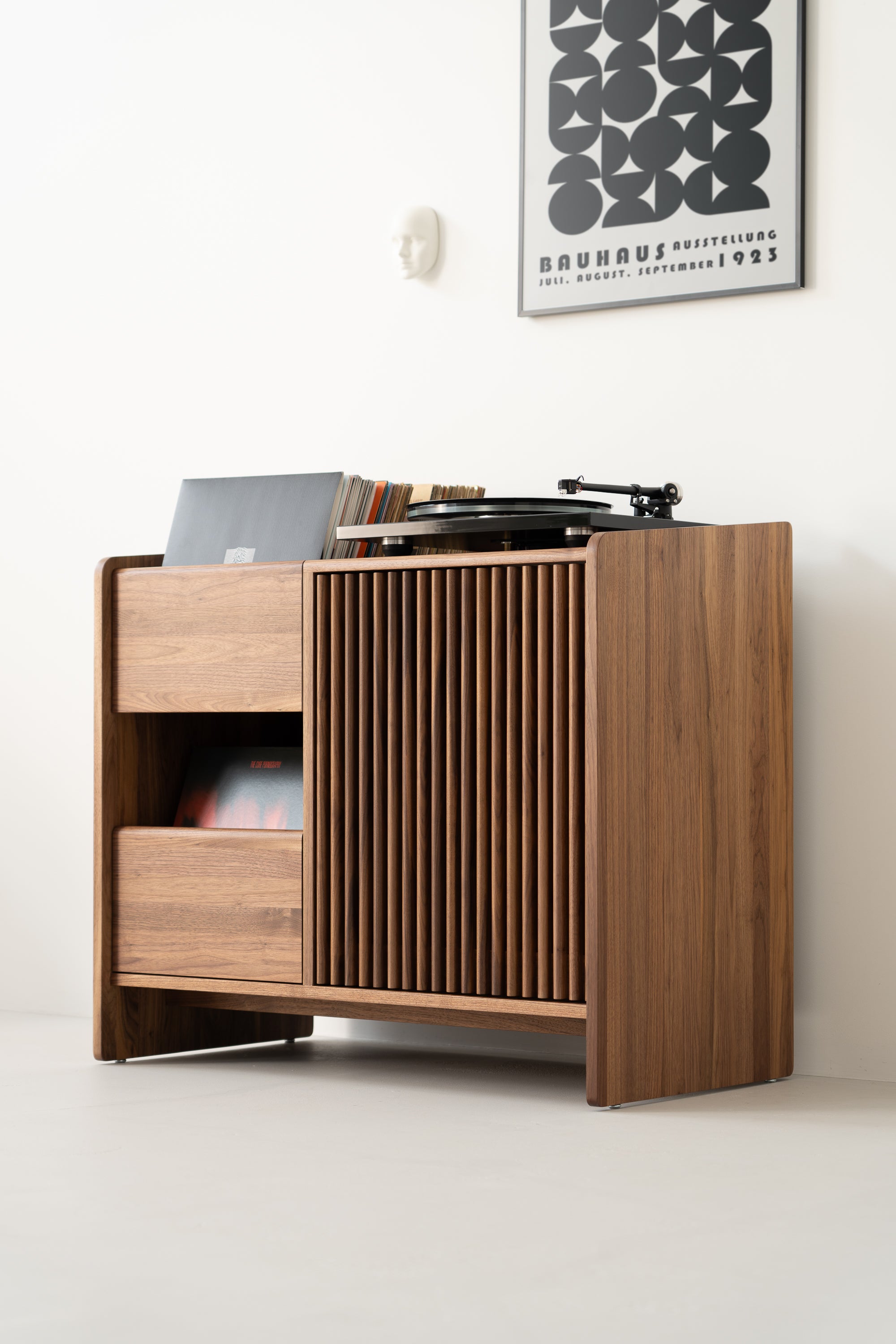 Wooden cabinet with vinyl records and a turntable, against a white wall with a framed Bauhaus poster above.