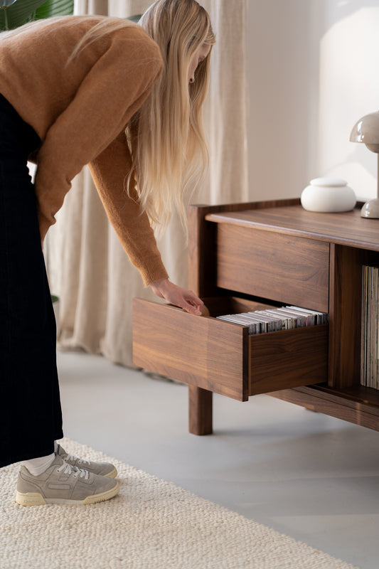 A person in a brown sweater and gray sneakers opens a drawer in a wooden cabinet filled with CDs.