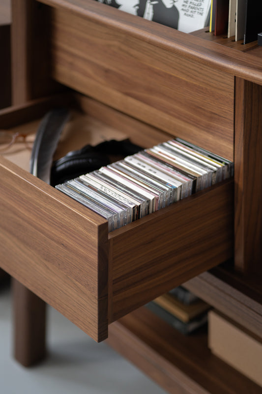 Wooden drawer partially open, showing a collection of CDs and a pair of headphones inside.