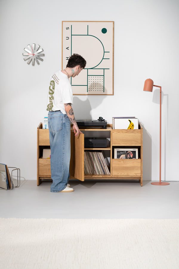 A man opens the slatted door of the FUSE X in solid oak, revealing an amplifier on a shelf and records below