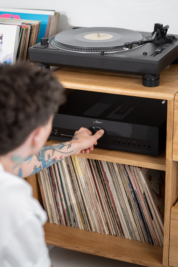 A man adjusts an amplifier in the FUSE X in solid oak, with records below and a turntable on top