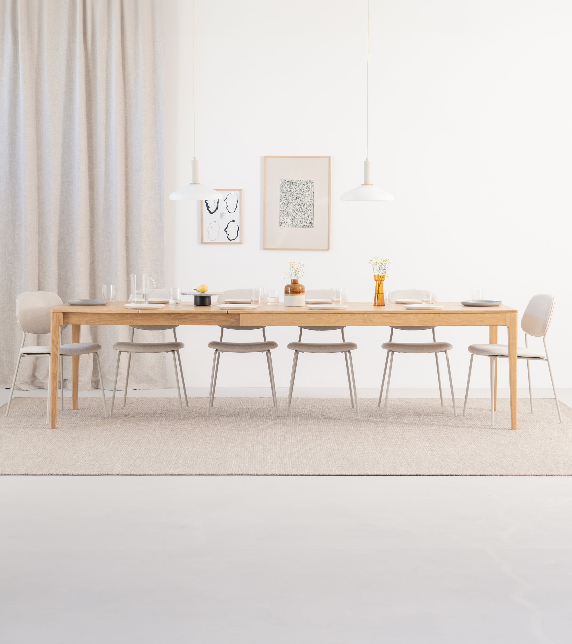 Minimalist dining room with a long wooden table, beige chairs, and two pendant lamps. Neutral tones and simple decor.