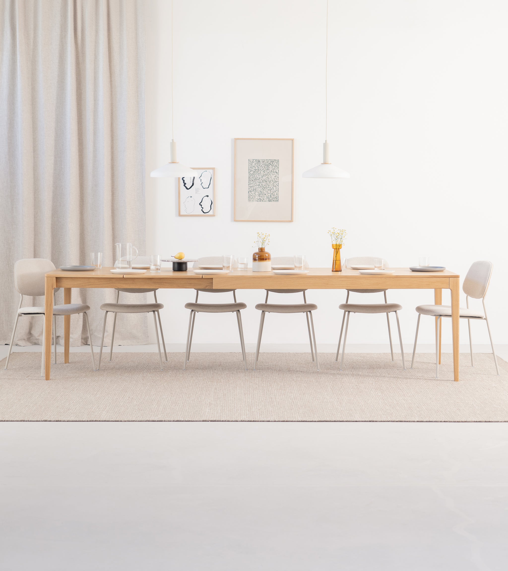 Minimalist dining room with a long wooden table, beige chairs, and two pendant lamps. Neutral tones and simple decor.