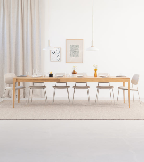 Minimalist dining room with a long wooden table, beige chairs, and two pendant lamps. Neutral tones and simple decor.