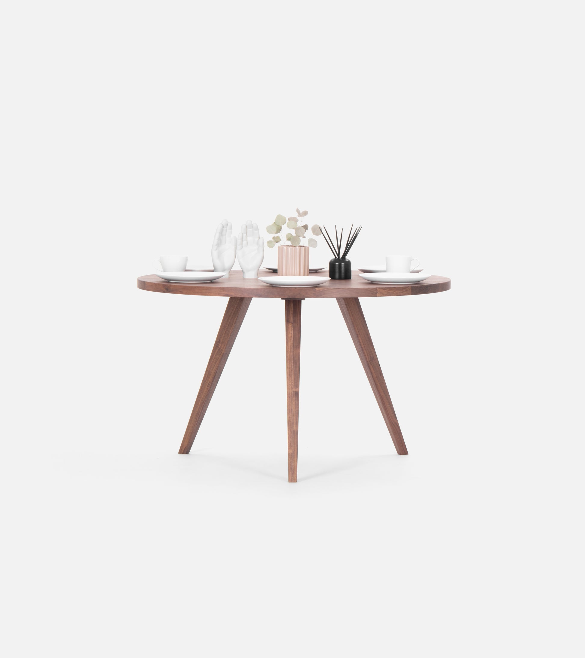 Round wooden table set for two, featuring cups, saucers, decorative white hands, a small plant, and a black reed diffuser.