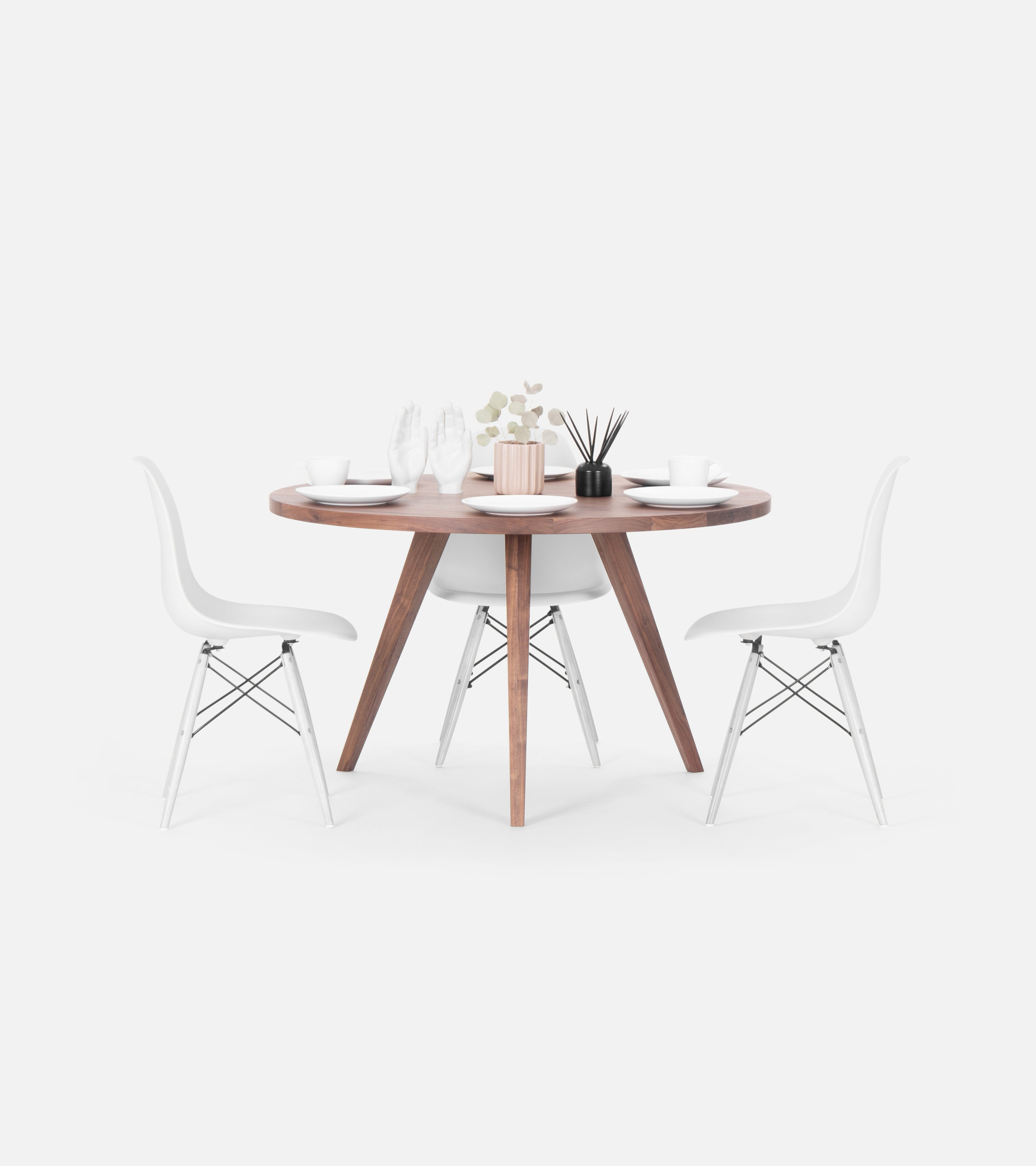 A round wooden table with minimalist decor, set with tableware, and flanked by two modern white chairs.