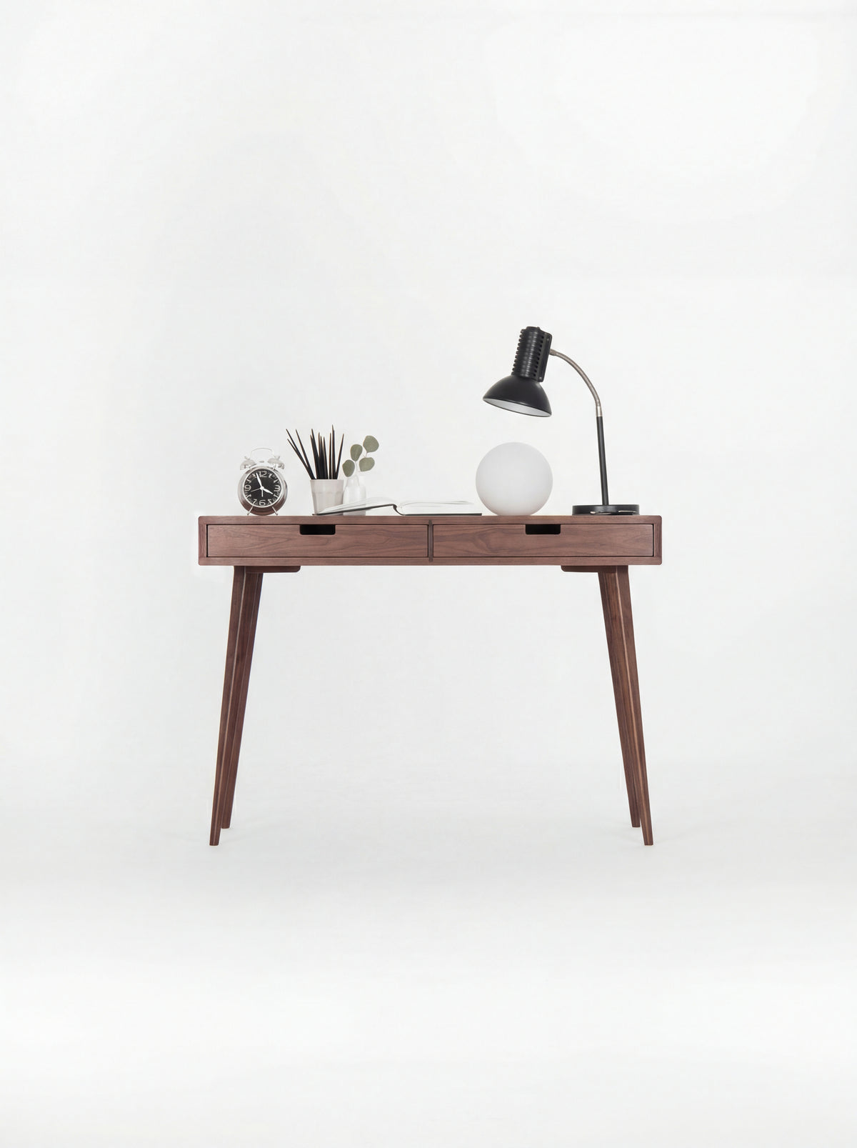 HEIM 105 desk in solid walnut, front view on a white background with lamps, an alarm clock and a pencil cup