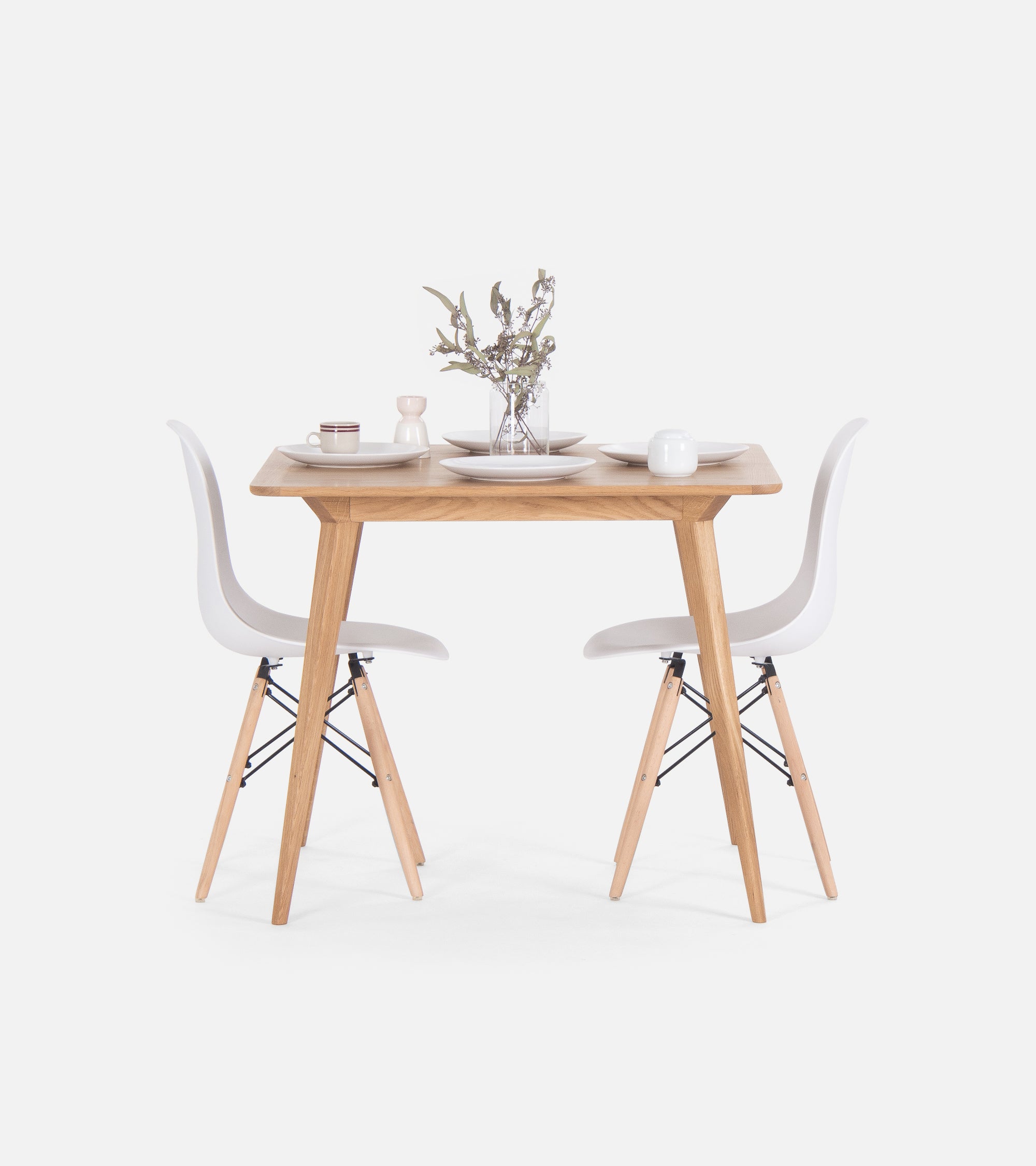 A minimalist wooden dining table with two white chairs, set for two with plates, cups, and a vase with flowers.