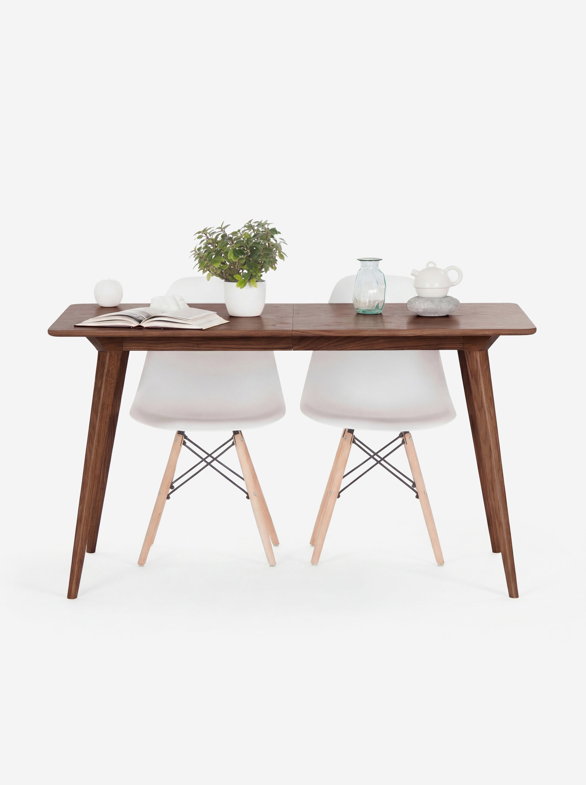 Wooden table with two white chairs, a plant, book, apple, vase, and teapot on top.