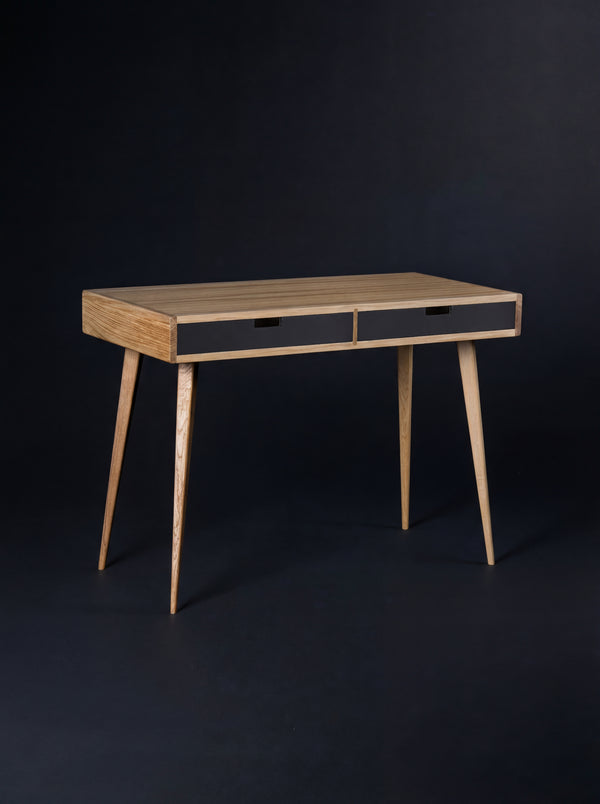 HEIM 105 desk in oak wood with two black drawer fronts, shown empty at an angle against a dark backdrop