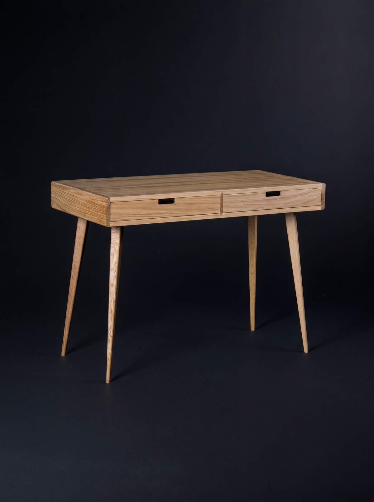 Empty HEIM 105 desk in oak wood on a dark background, angled to show wooden drawer fronts and splayed legs