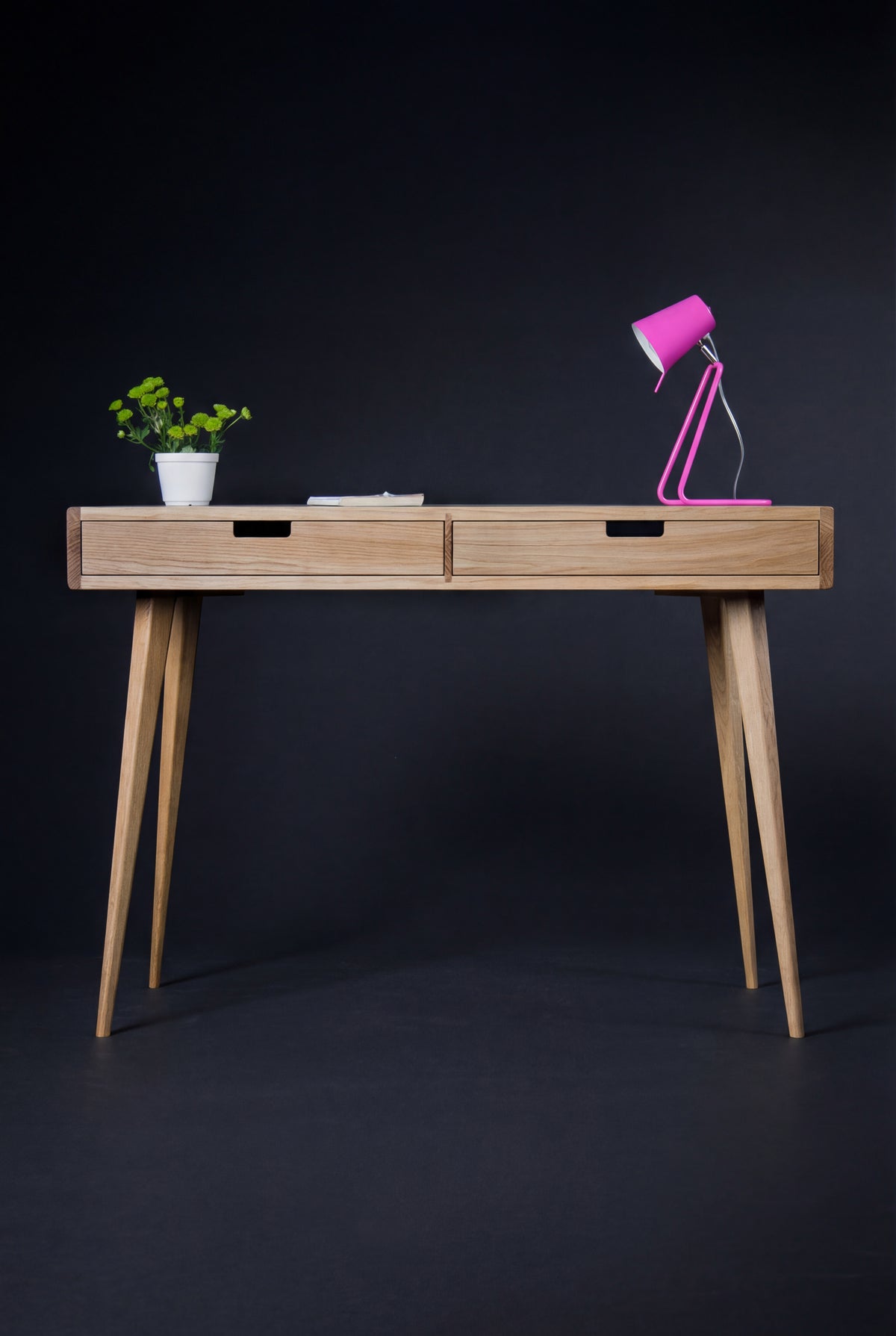 Front view of the HEIM 105 desk in oak wood with wooden drawer fronts, a pink lamp, a plant and a thin book