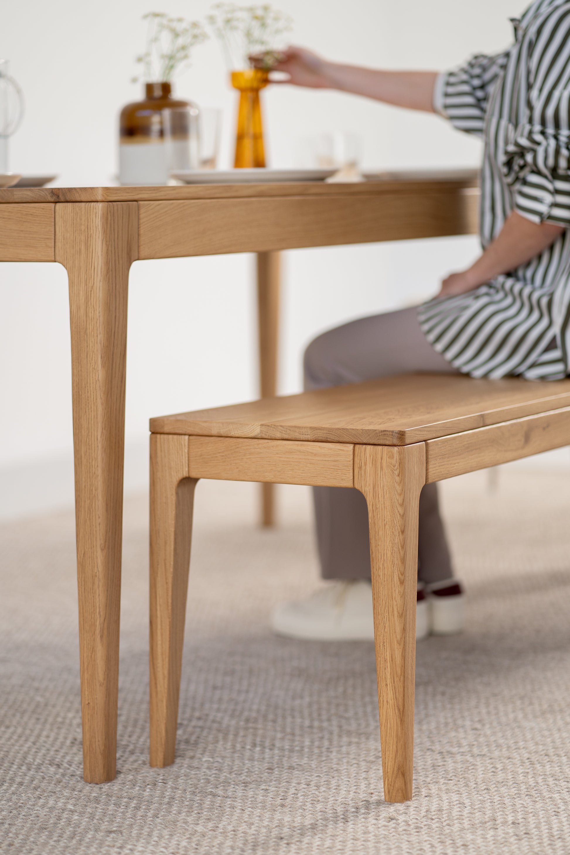 Wooden dining set with bench, person adjusting flowers in amber vase on table.