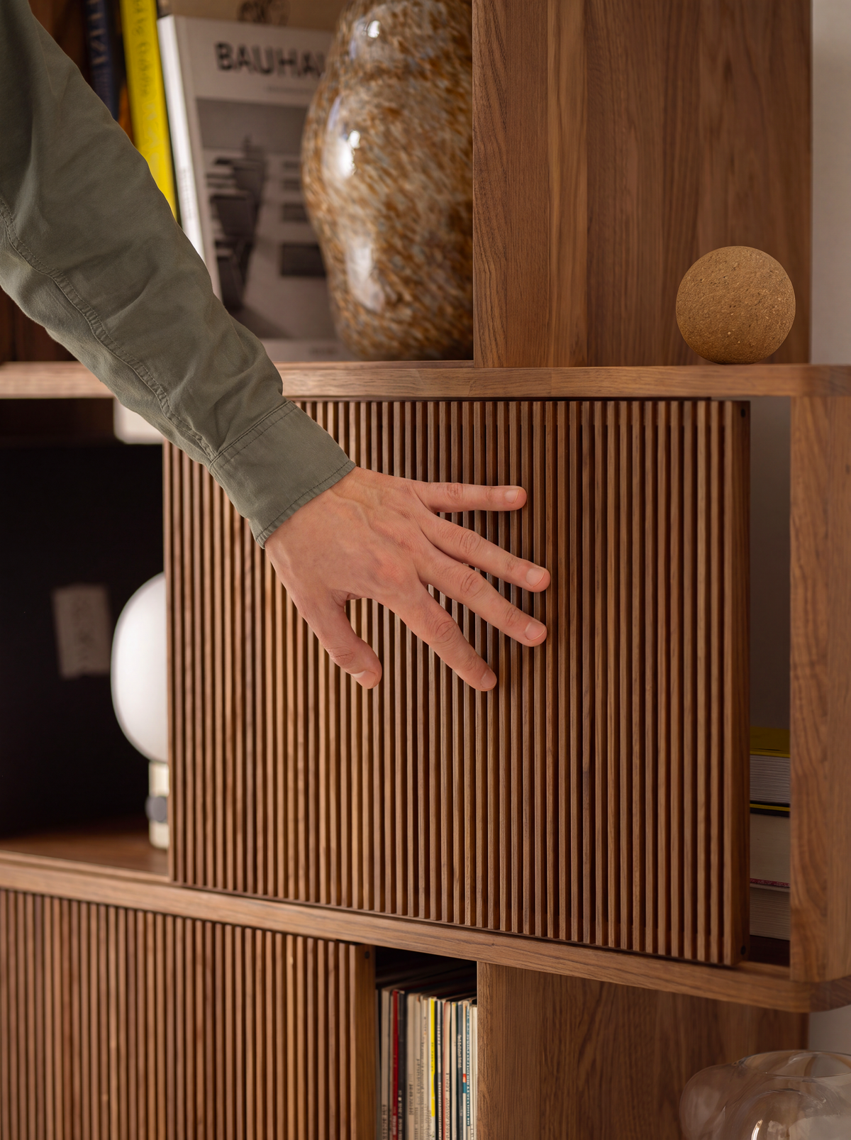 A hand moves a slatted front along a shelf of MOLL DUO in solid walnut, beneath books and a glass vase