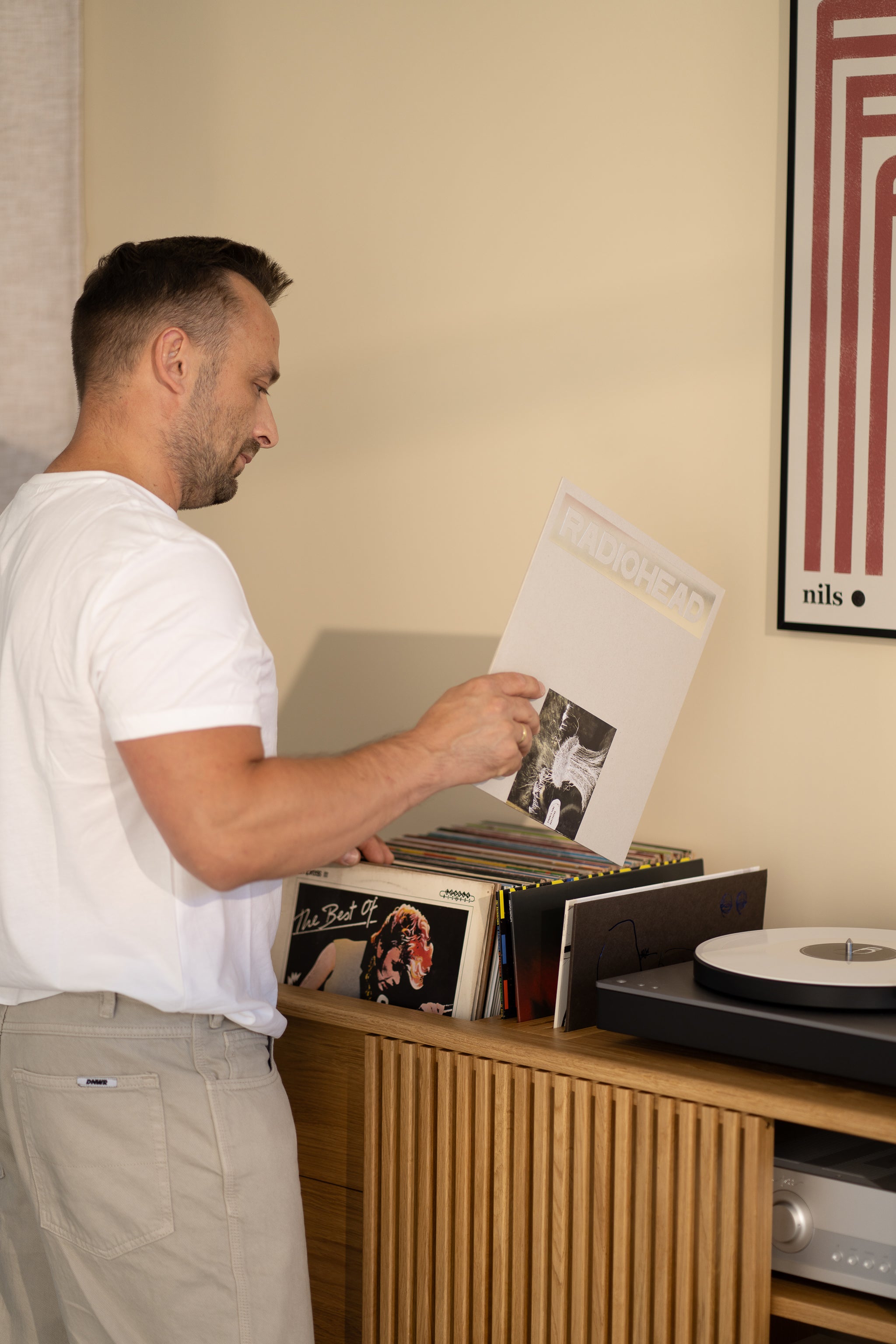 Man in white shirt browsing records next to a turntable on a wooden cabinet.