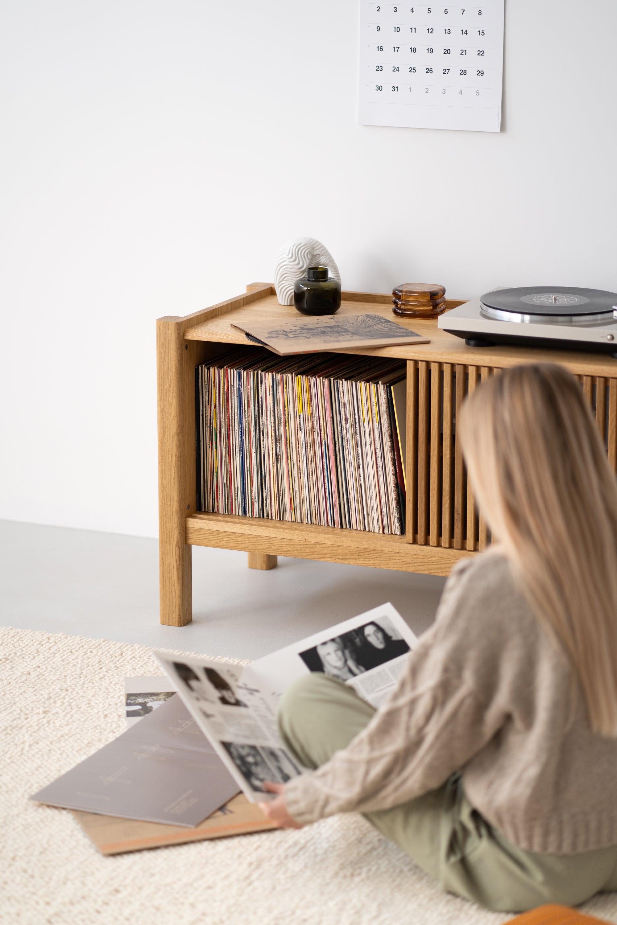 Person sitting on the floor looking at vinyl records near a wooden shelf with more records and a turntable.