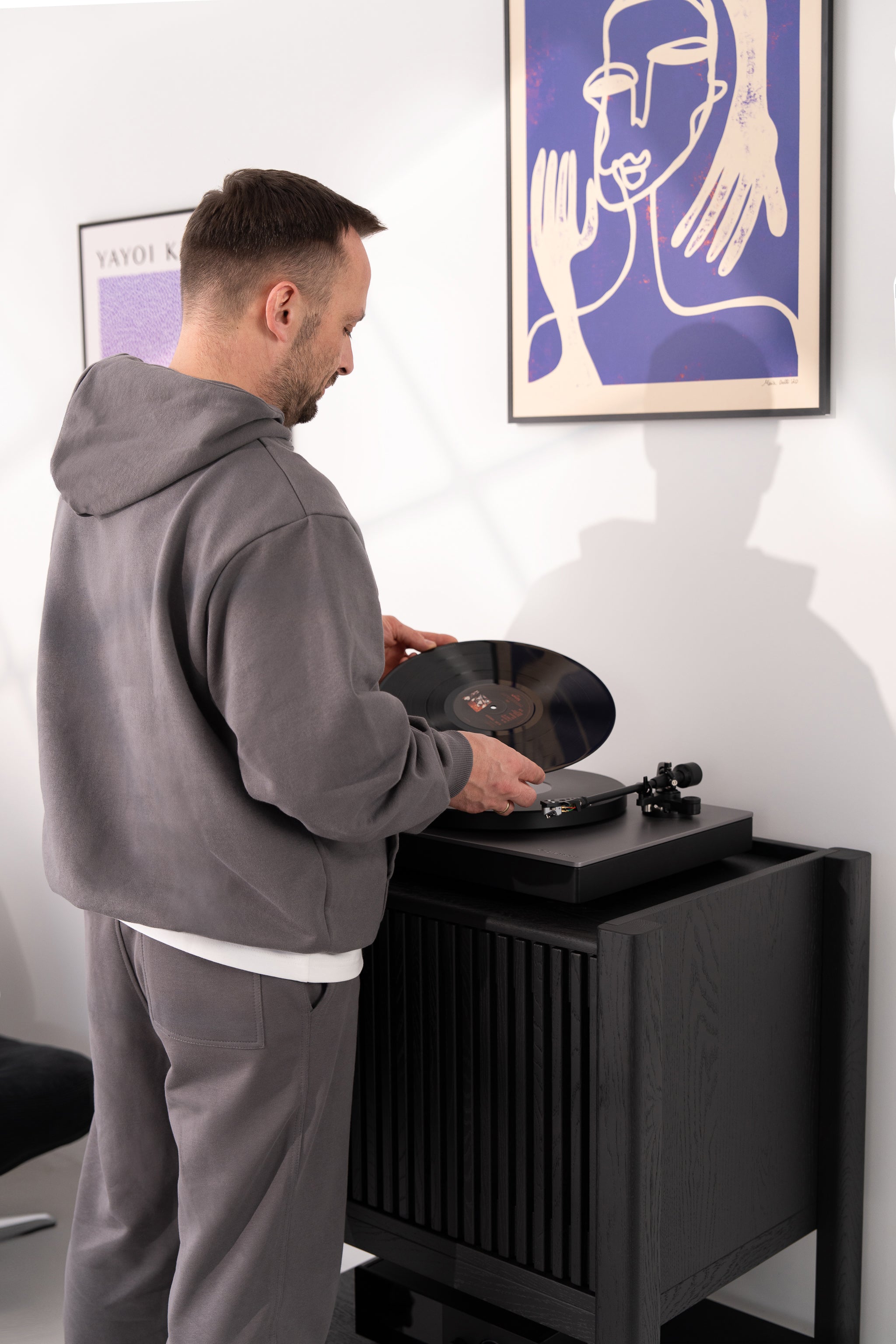 Man in a gray hoodie placing a vinyl record on a turntable beneath modern artwork.