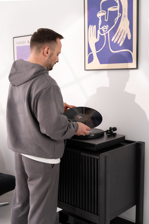 Man in a gray hoodie placing a vinyl record on a turntable beneath modern artwork.