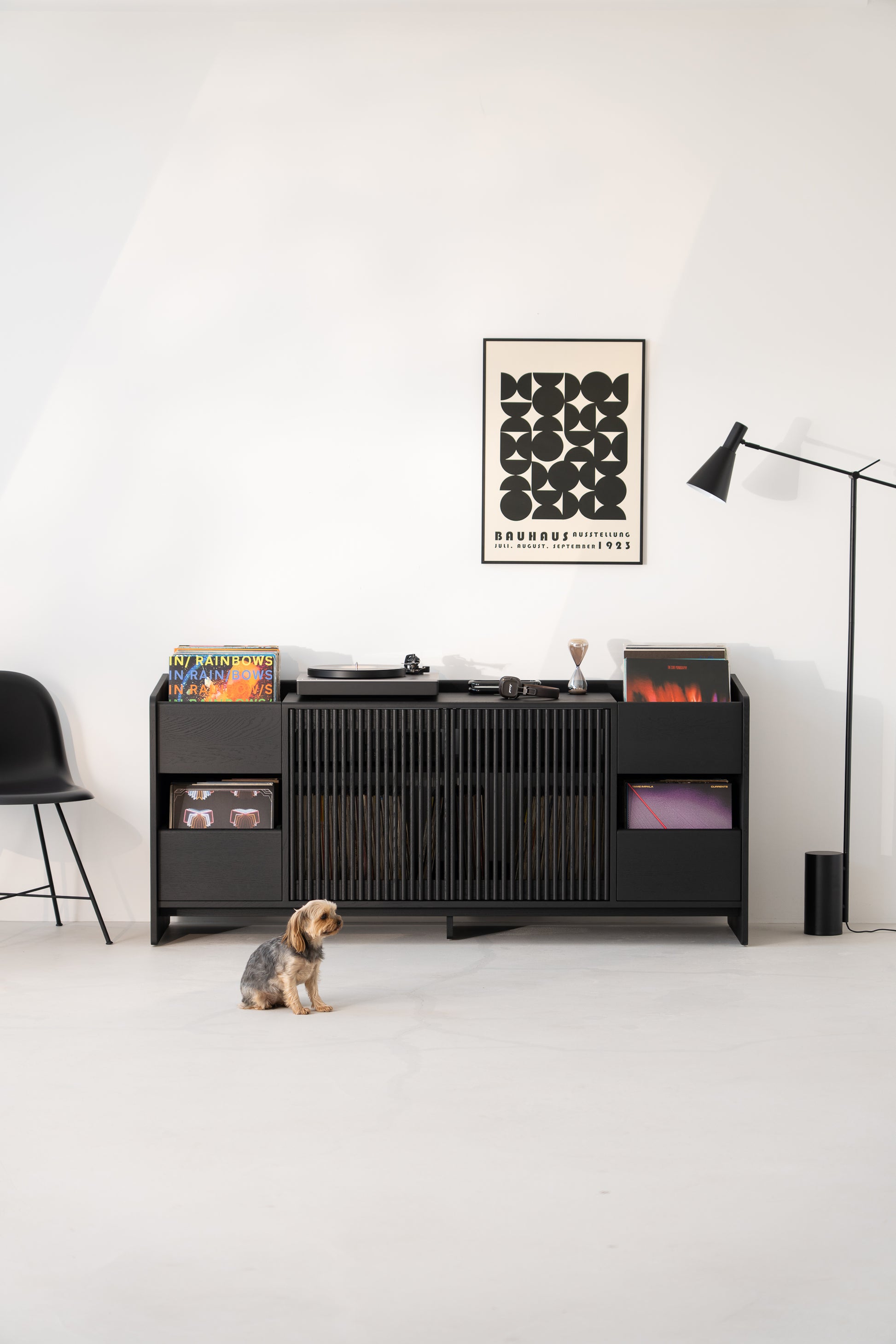 Minimalist room with a black sideboard, turntable, records, Bauhaus poster, and a small dog on the floor.