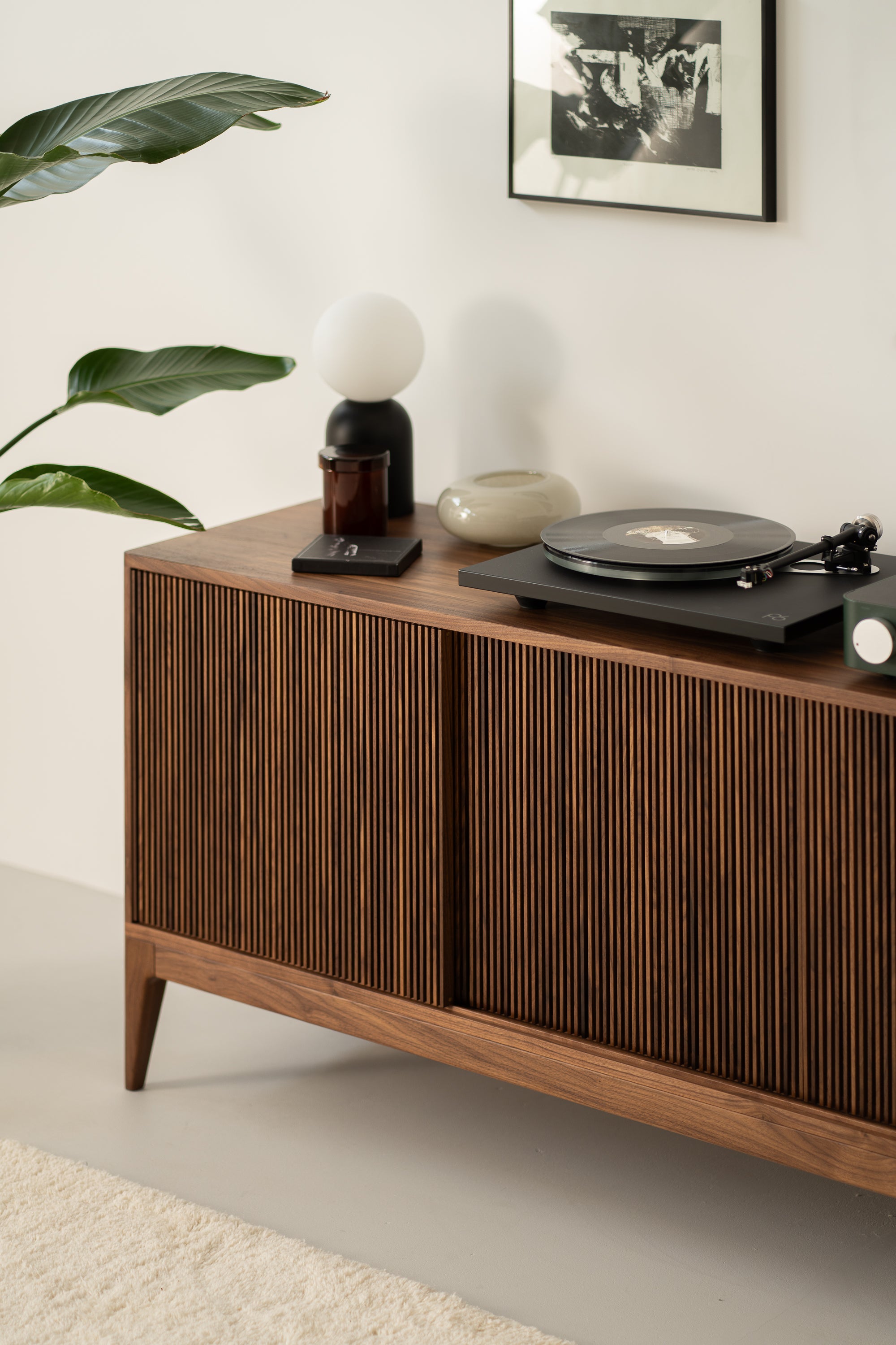 Wooden cabinet with a record player, lamp, and small items on top. Plant on the left, framed photo on white wall above.
