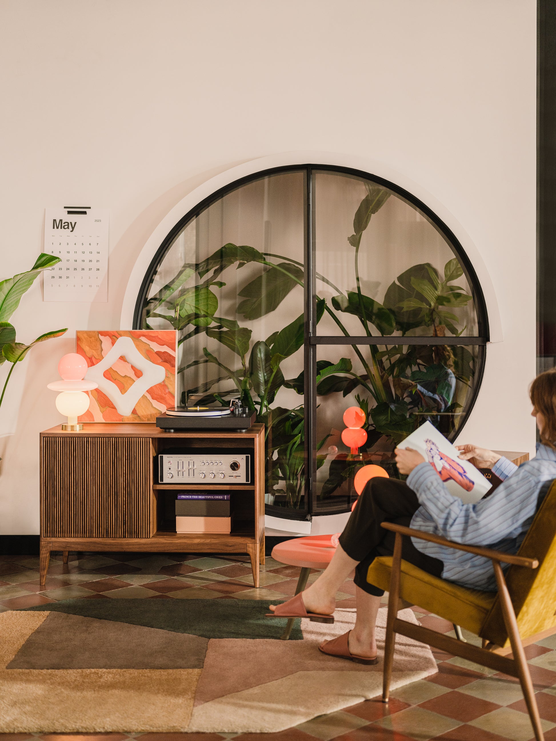 Cozy room with plants, a circular window, a calendar, and a person reading a magazine beside a vintage cabinet.