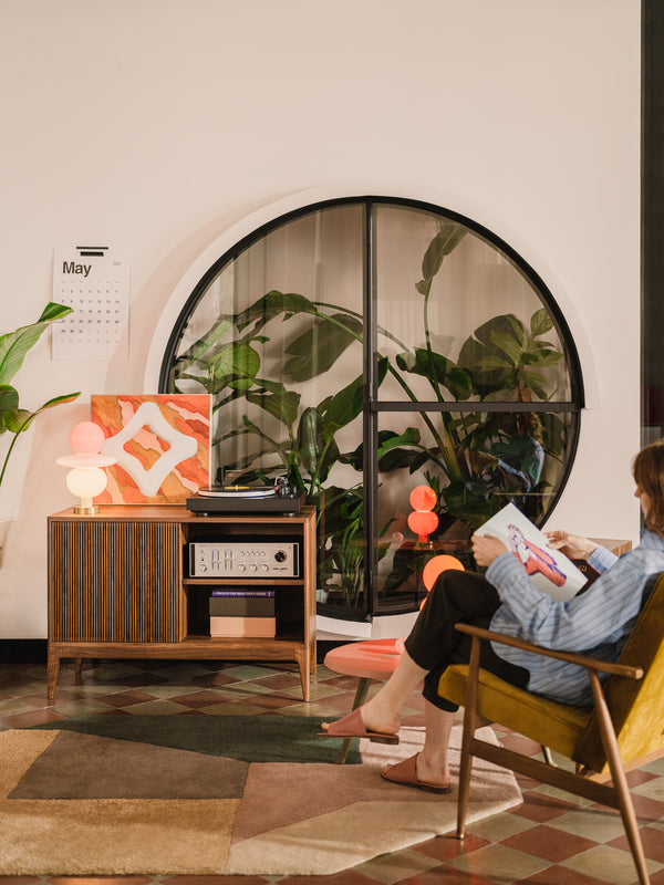 Cozy room with plants, a circular window, a calendar, and a person reading a magazine beside a vintage cabinet.