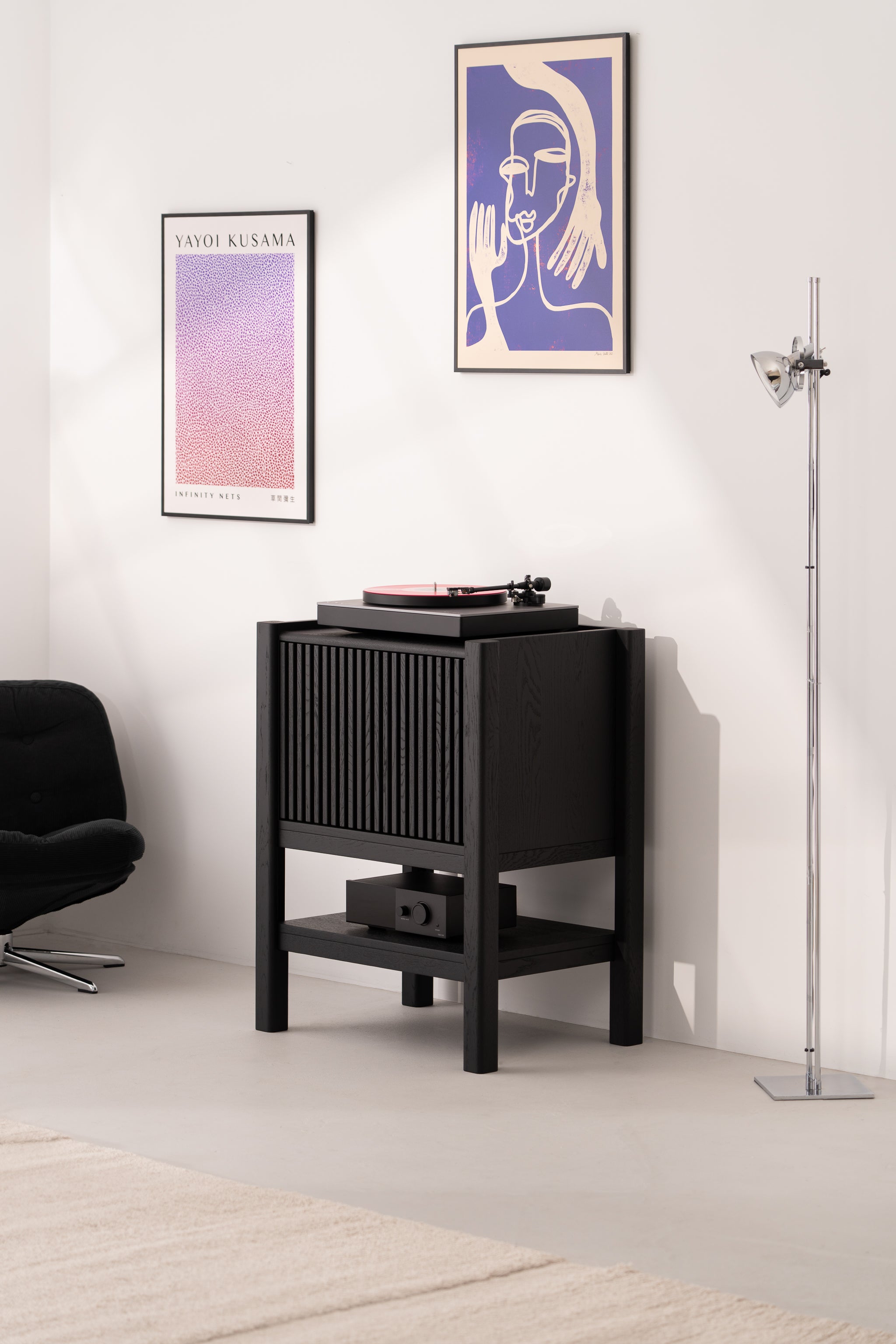 Room with a vinyl player on a black stand, abstract wall art, a floor lamp, and a black chair.