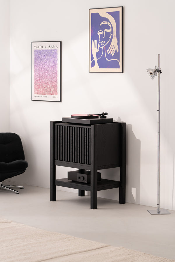 Room with a vinyl player on a black stand, abstract wall art, a floor lamp, and a black chair.