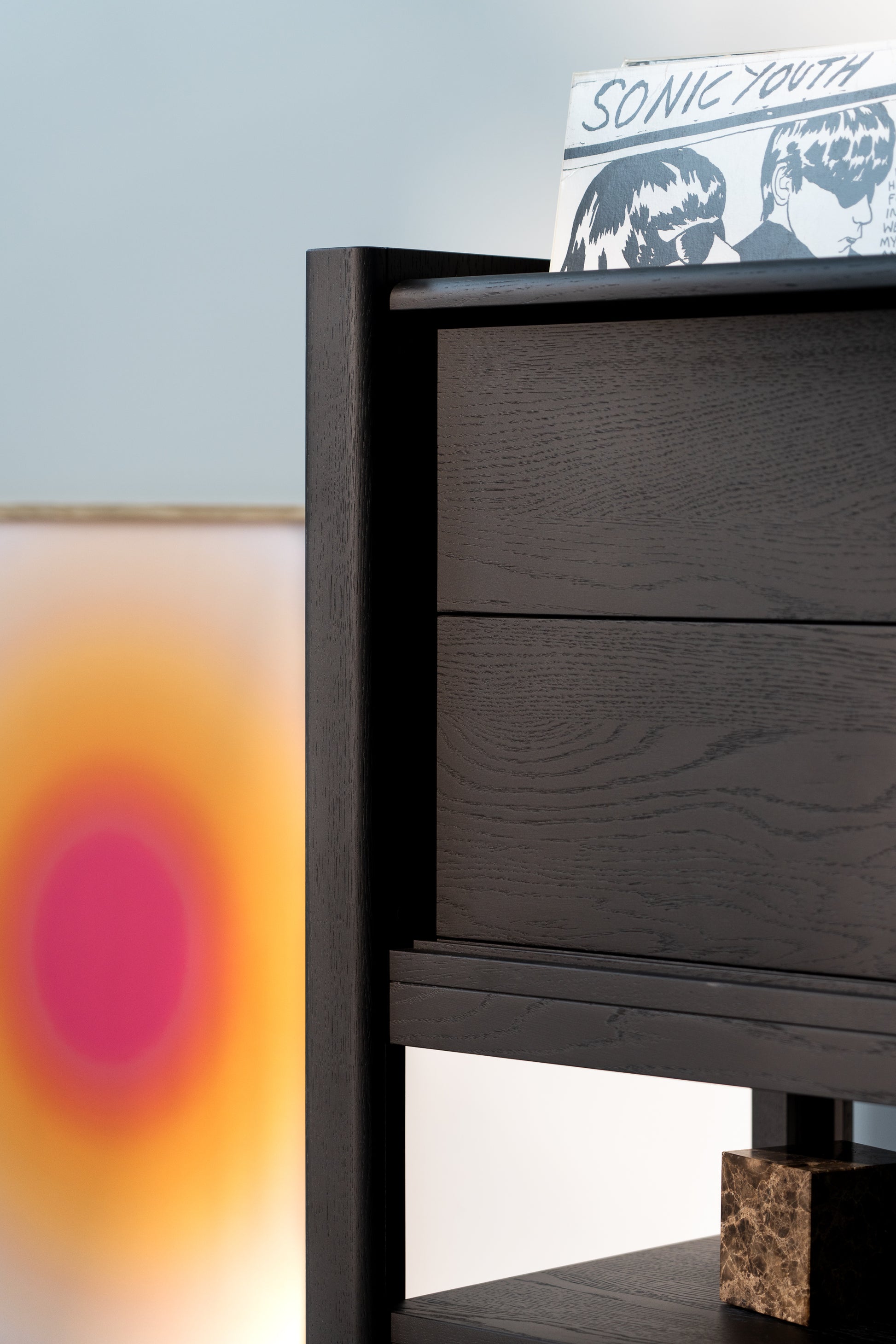 Dark wooden shelf with a vinyl record on top. A blurred artwork with a pink and orange gradient is in the background.
