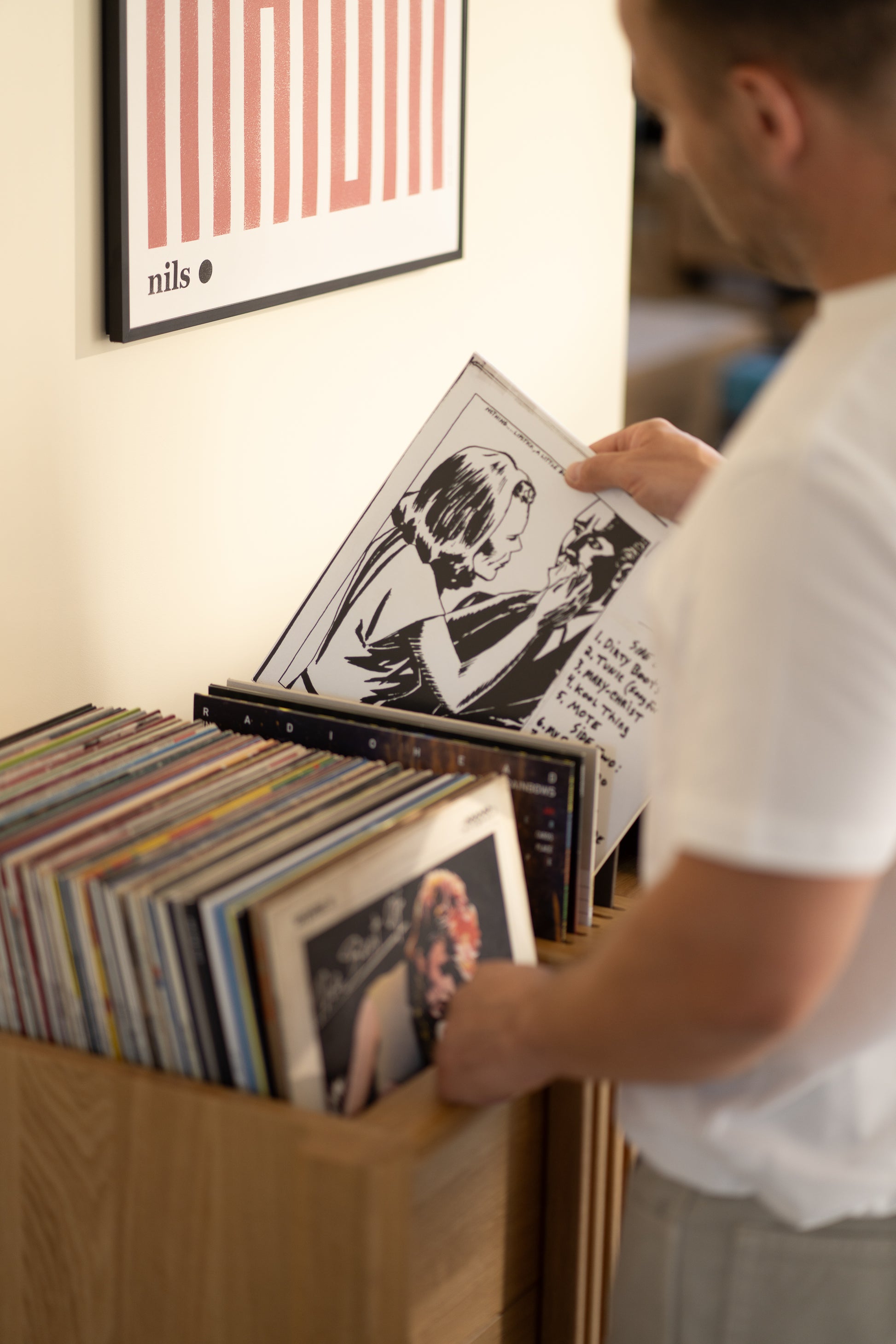Person browsing vinyl records in a wooden shelf beneath a wall poster.