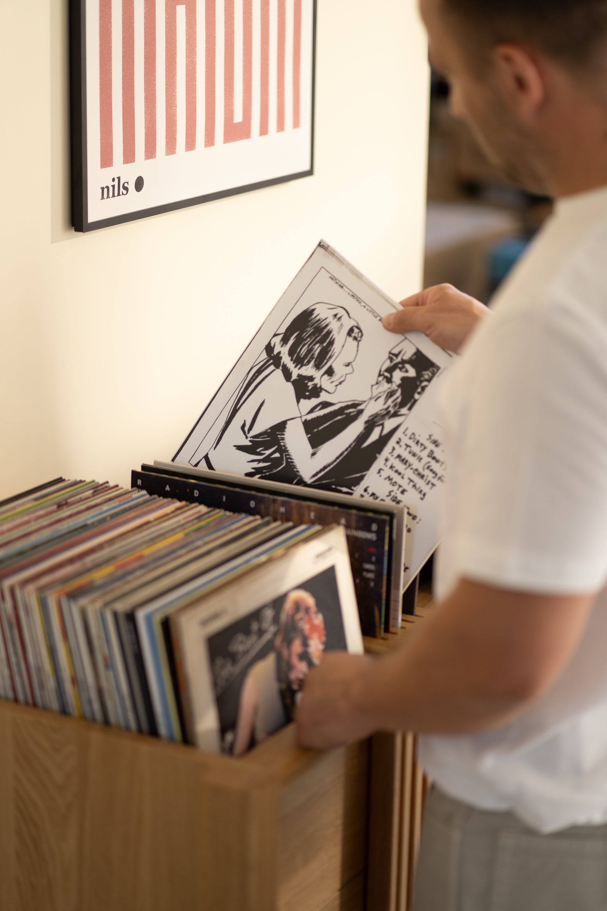 Person browsing vinyl records in a wooden shelf beneath a wall poster.
