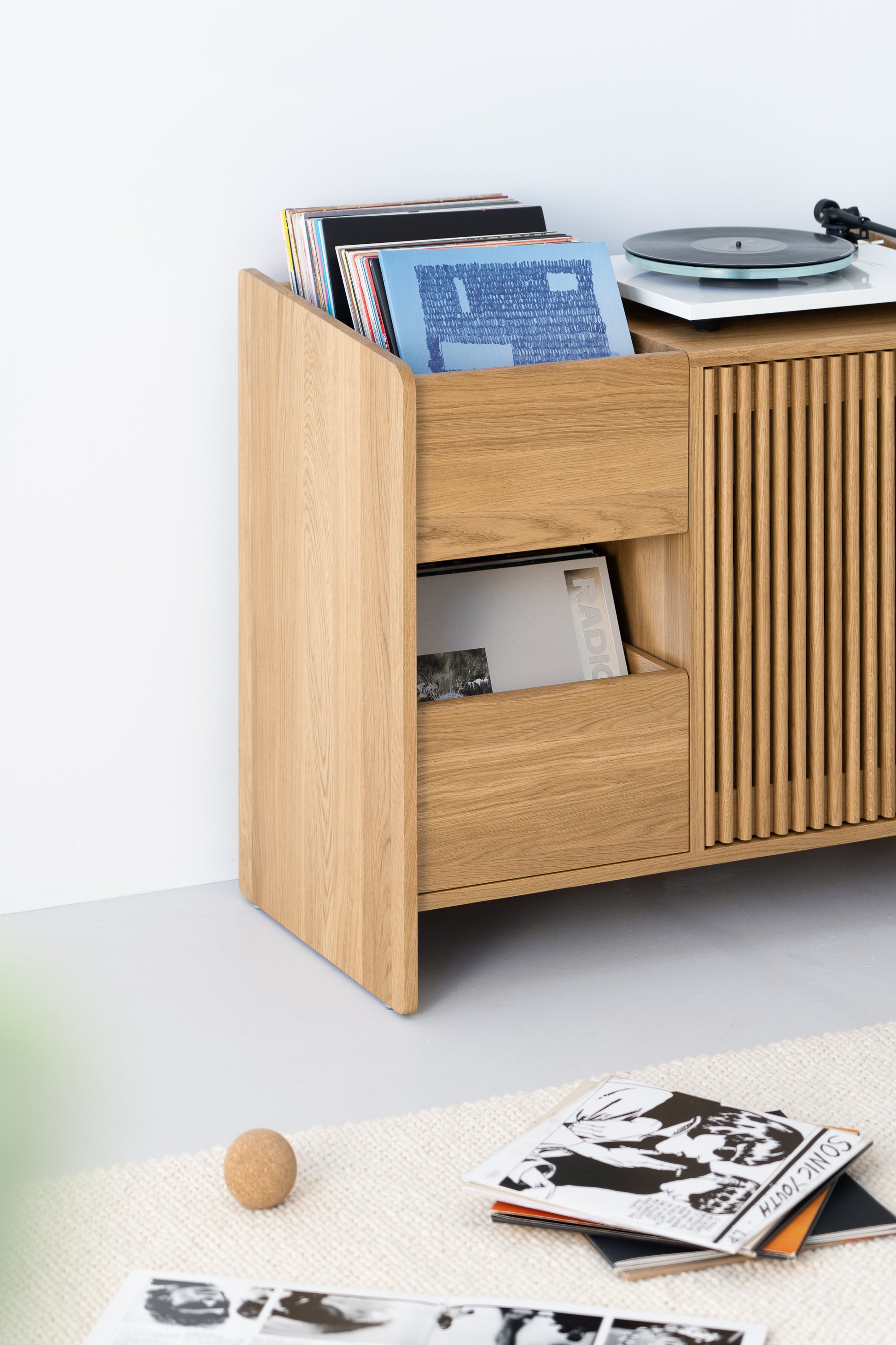 Wooden record cabinet with vinyl records and turntable. Floor scattered with opened vinyl covers and a ball.