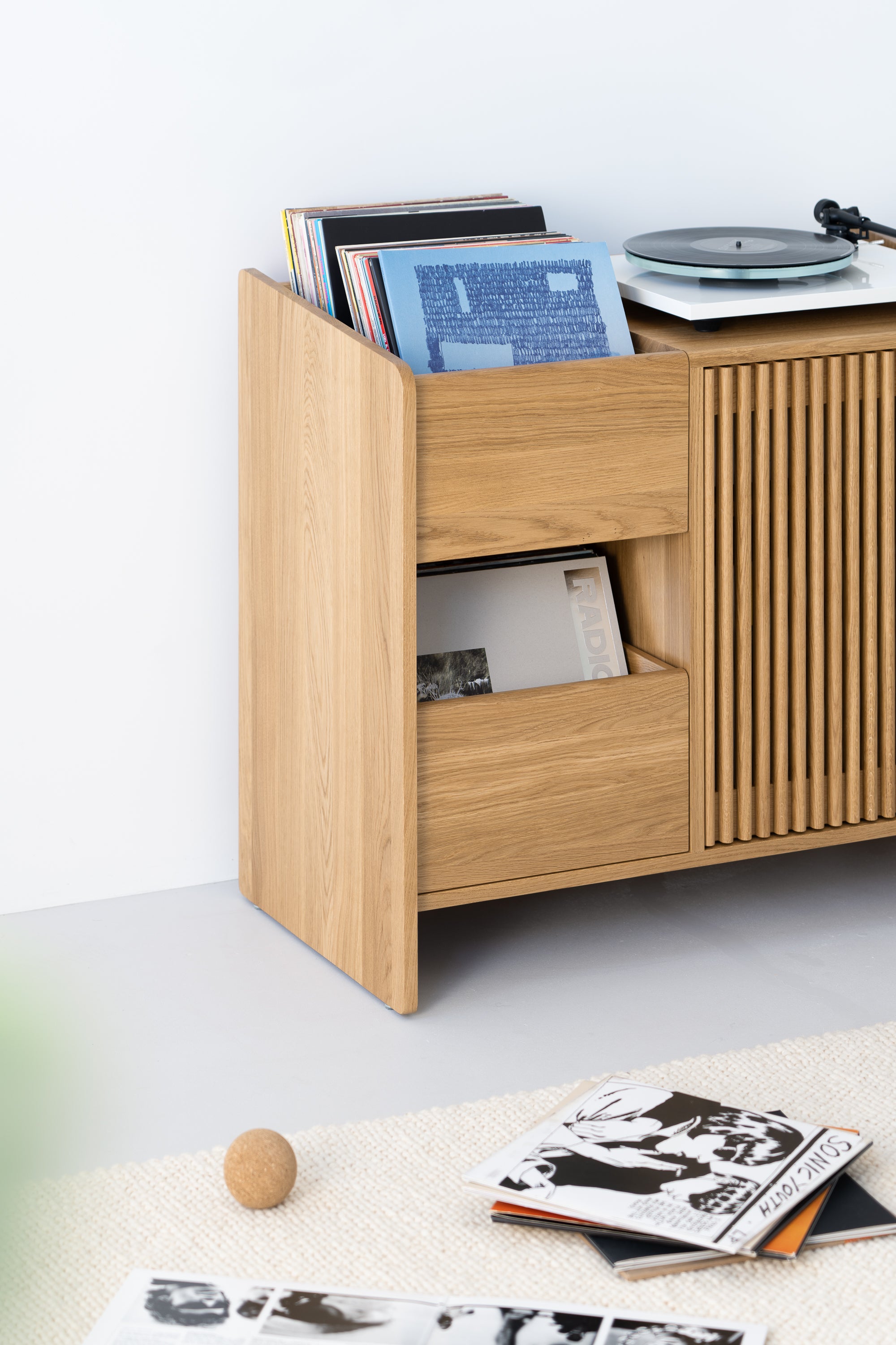 Wooden record cabinet with vinyl records and turntable. Floor scattered with opened vinyl covers and a ball.