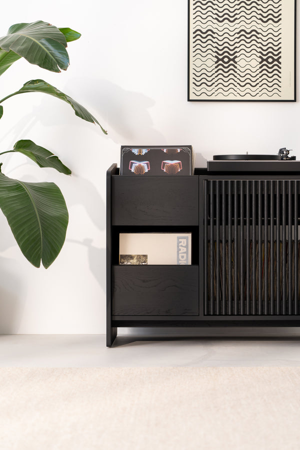 Vinyl records and a record player in a modern black cabinet, with a leafy plant and abstract art on the wall.