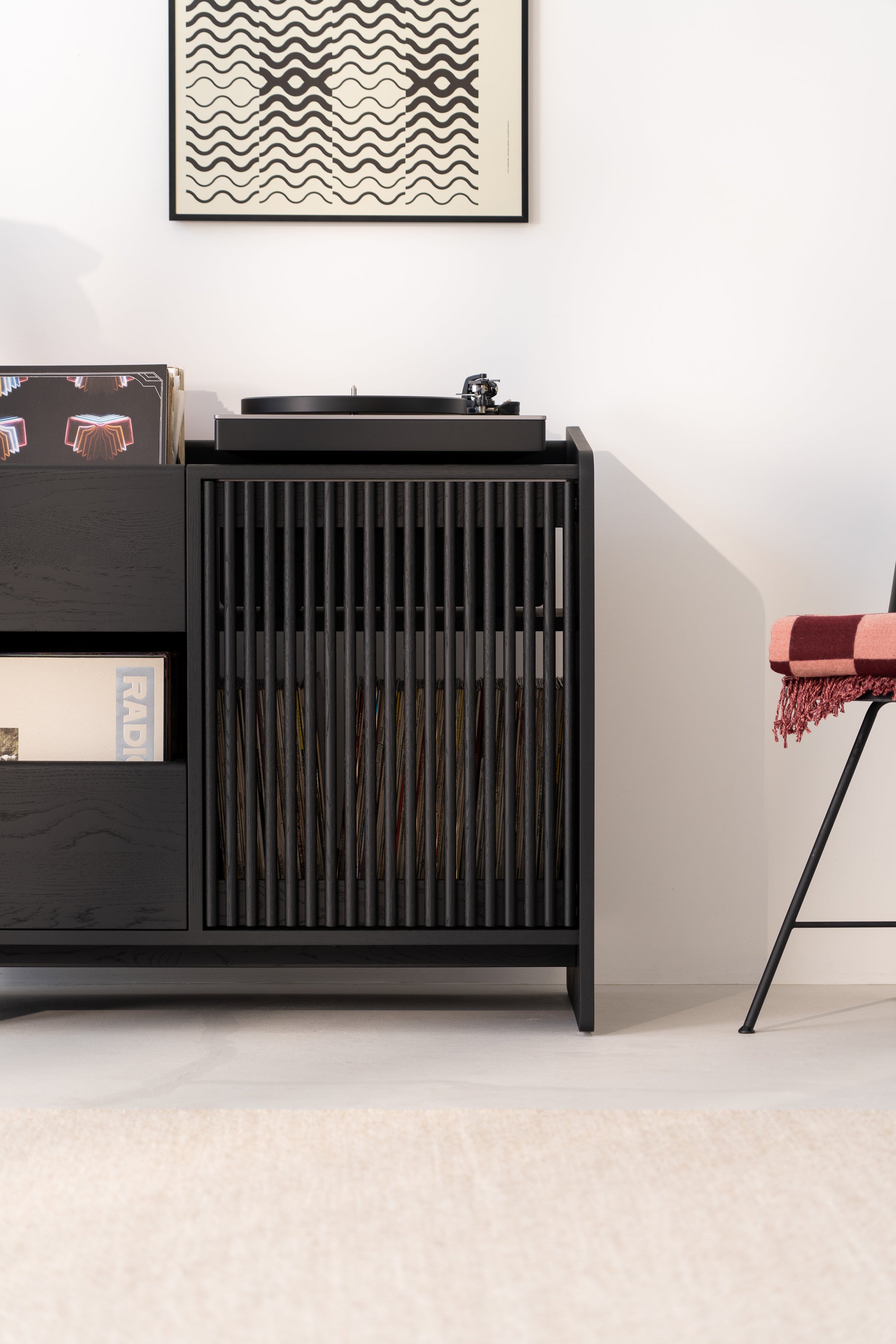 Vinyl record player on a sleek black cabinet with records, next to a chair. Abstract art hangs above.