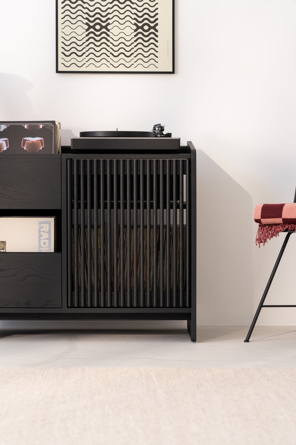 Vinyl record player on a sleek black cabinet with records, next to a chair. Abstract art hangs above.