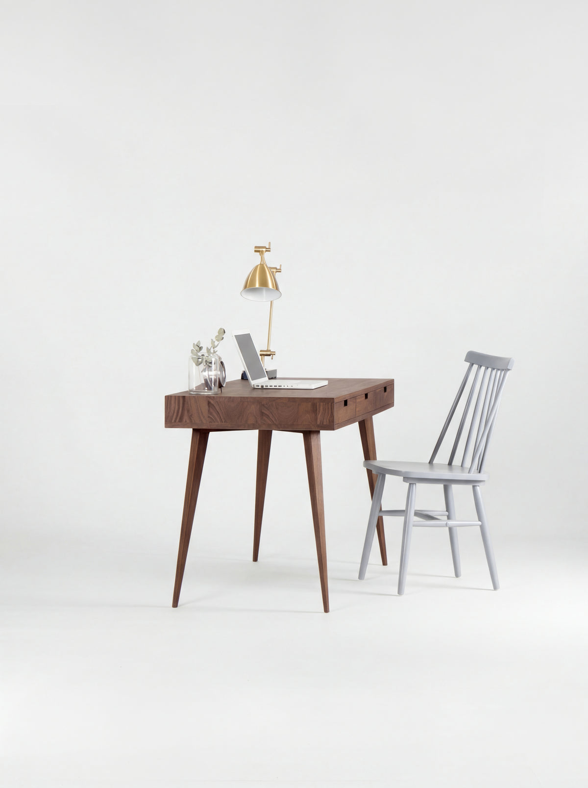 HEIM 140 solid walnut computer desk seen from the short end, with a grey spindle chair, laptop and brass lamp