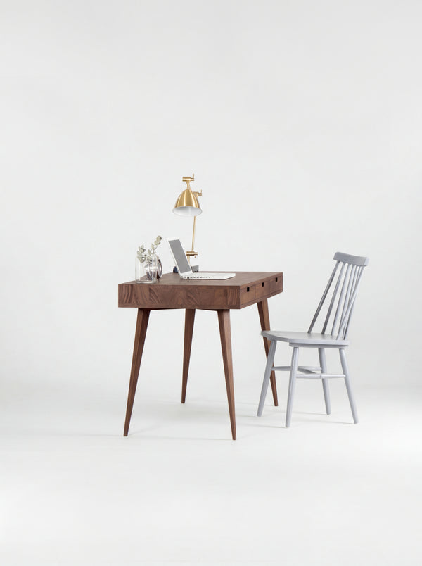 HEIM 140 solid walnut computer desk seen from the short end, with a grey spindle chair, laptop and brass lamp