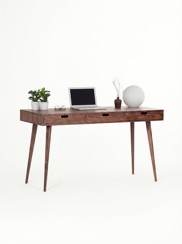 HEIM 140 solid walnut desk at a three-quarter angle with a laptop, plants and a globe lamp