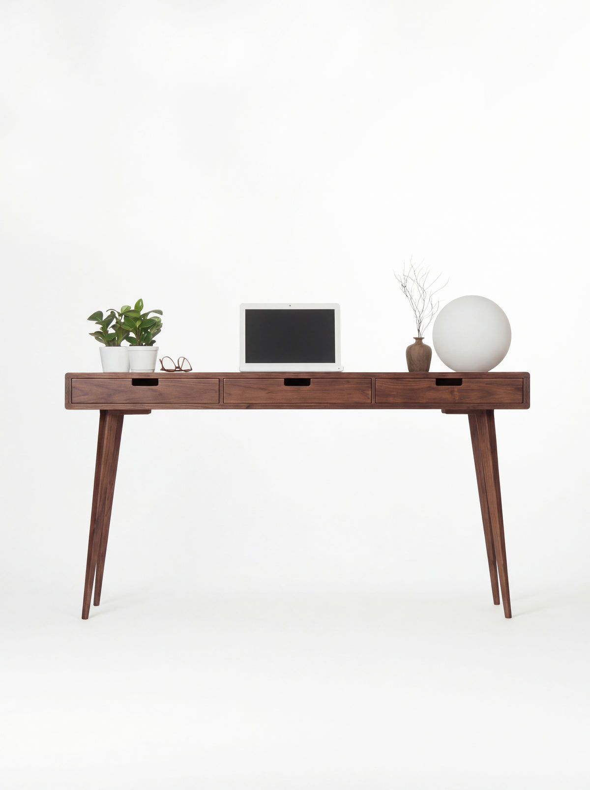 Front view of the HEIM 140 desk in solid walnut, with splayed legs, a laptop and a globe lamp