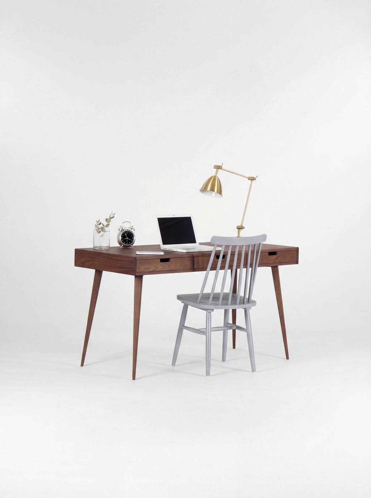 HEIM 140 walnut wood desk with a grey spindle chair pulled up, a laptop, alarm clock and brass lamp on top