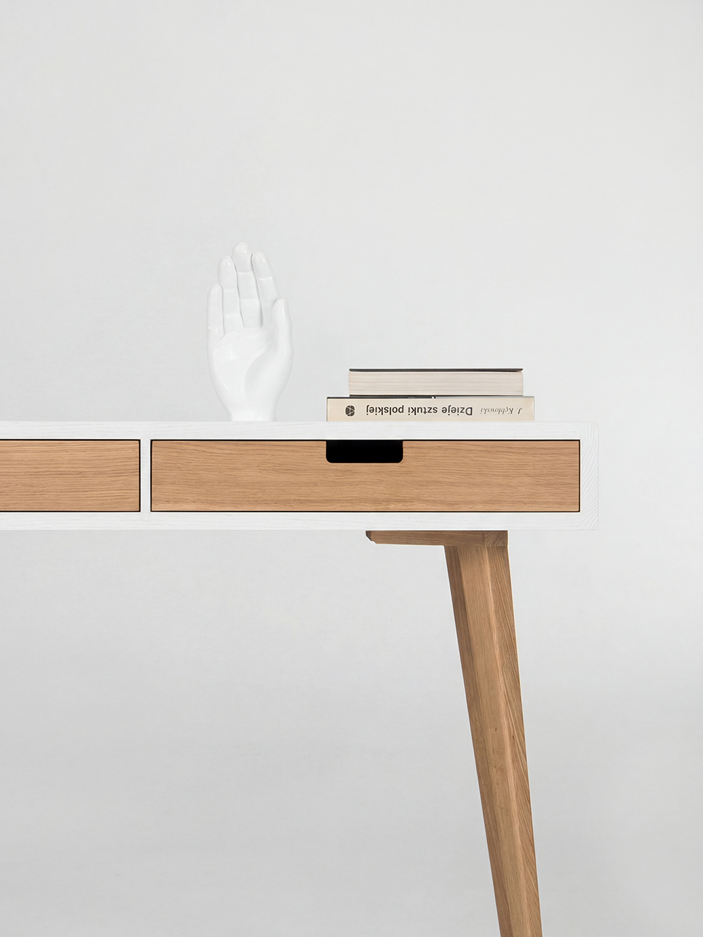 Books and a white hand sculpture on the right end of the HEIM 140 desk in solid oak, above an angled leg