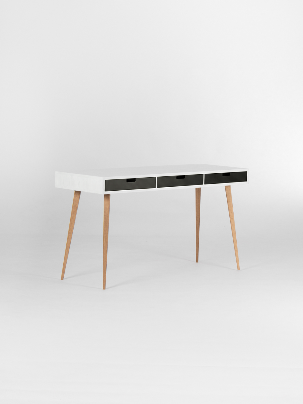 Angled view of the HEIM 140 desk in solid oak, with an empty white top, three black drawer fronts and oak legs
