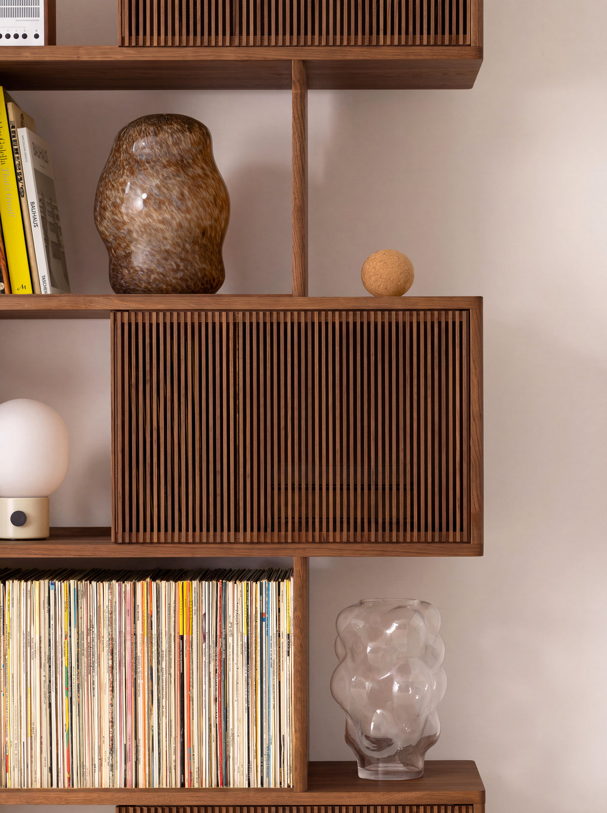 MOLL in solid walnut, close-up of a slatted front between shelves holding records, a glass vase and a table lamp